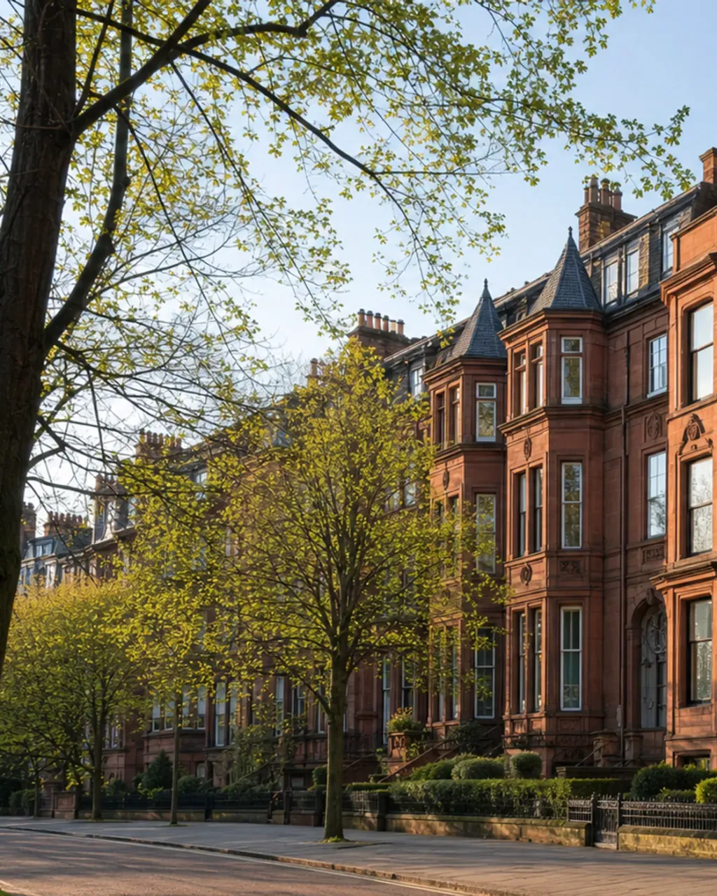 Red sandstone tenement bay windows and turrets behind trees in early leaf on a quiet Hyndland street