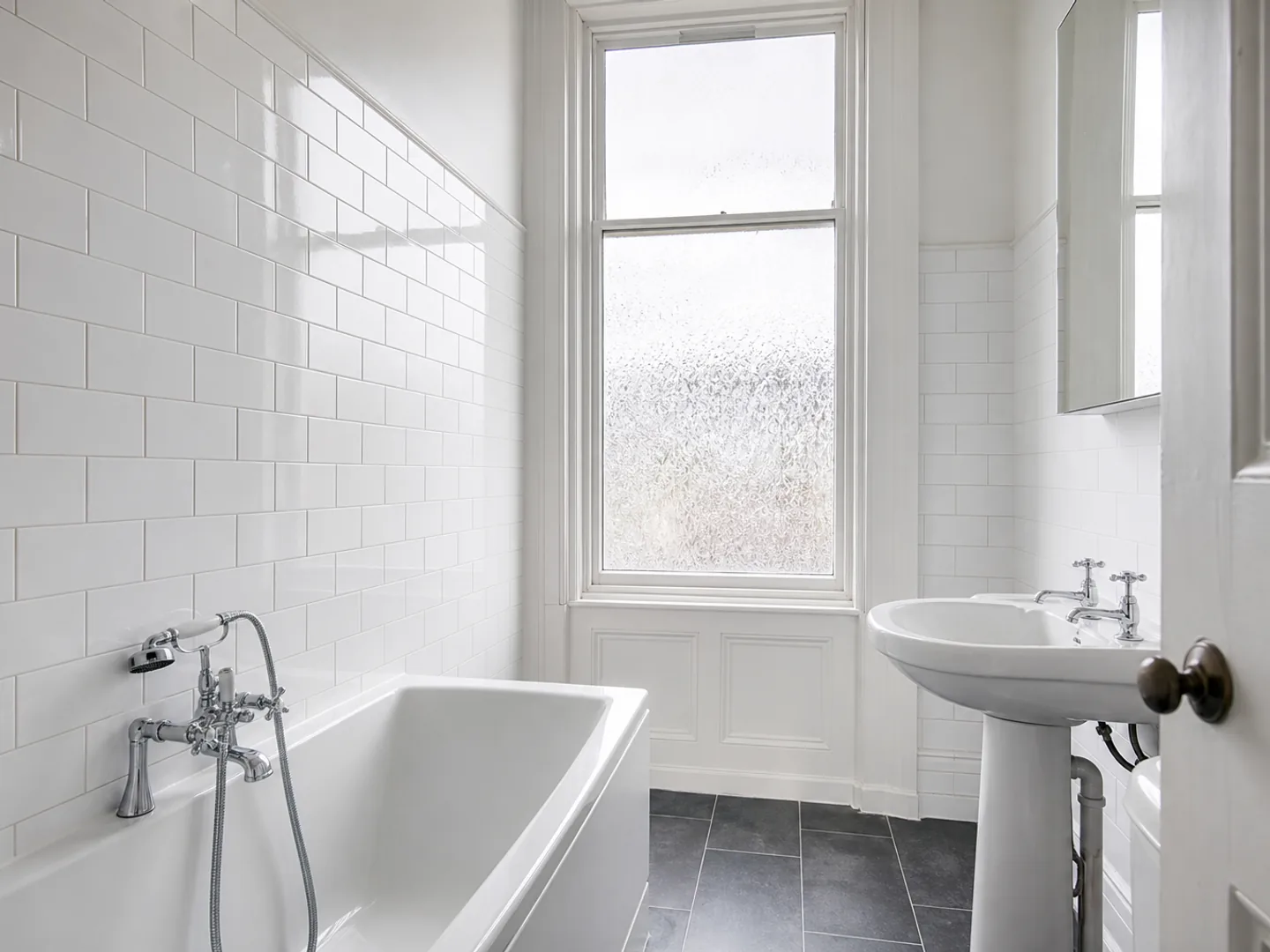 A white tiled tenement bathroom with a bath, pedestal basin and slate floor, cleaned and empty