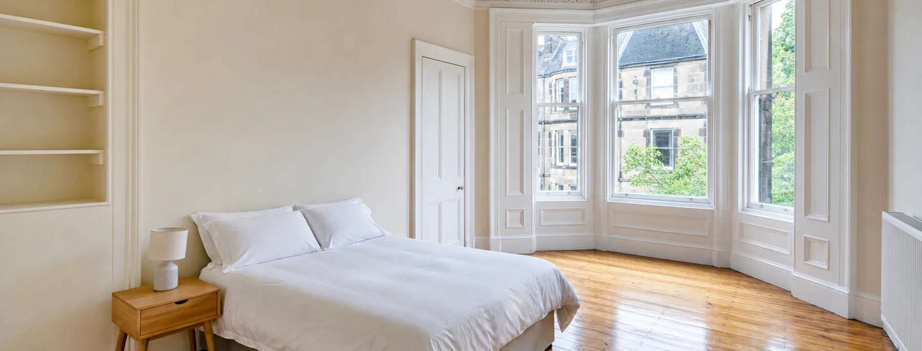 A cream tenement bedroom with a bare double bed, stripped floorboards and a bay window over the street